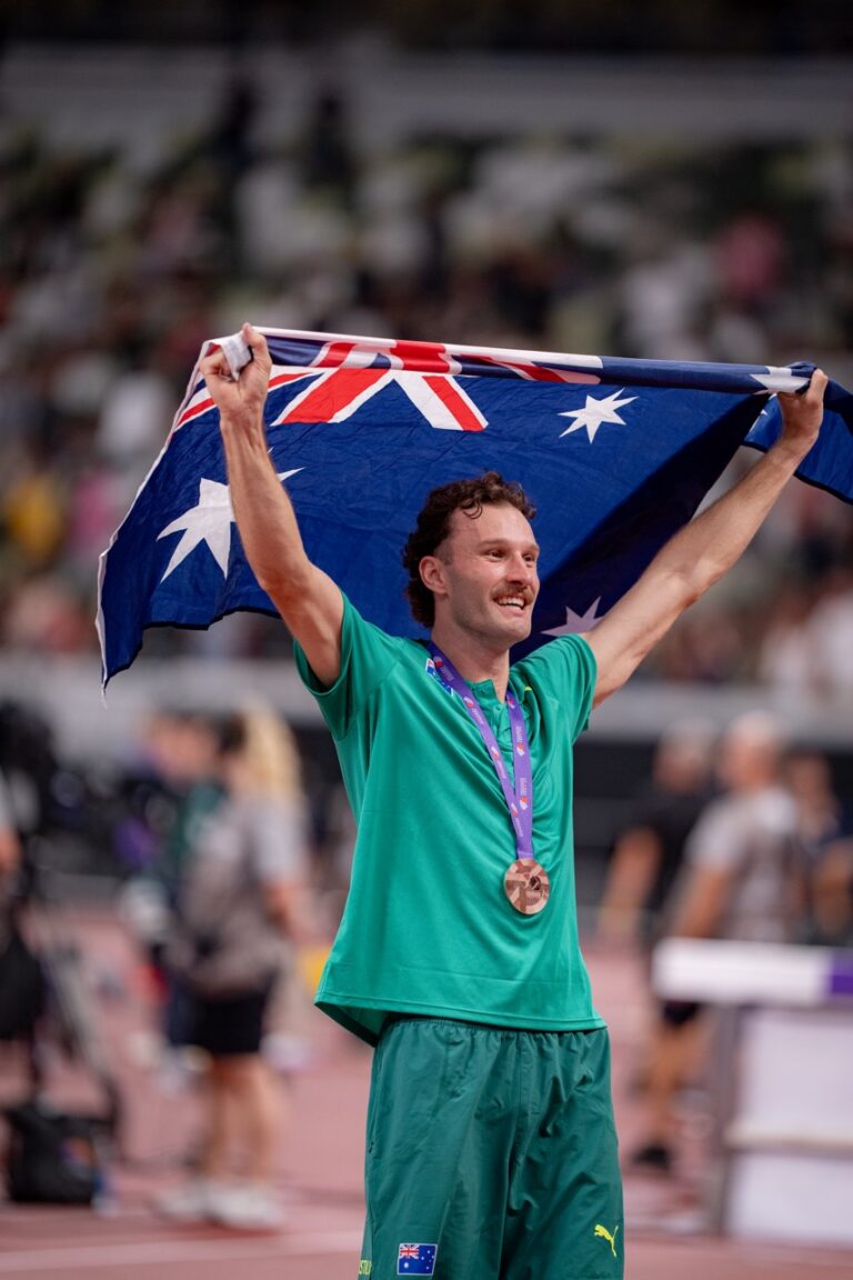 Athlete on a track raises the Australian flag above his head, wearing a green uniform and a bronze medal around his neck.