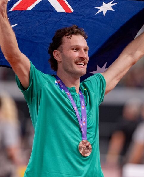 Athlete on a track raises the Australian flag above his head, wearing a green uniform and a bronze medal around his neck.