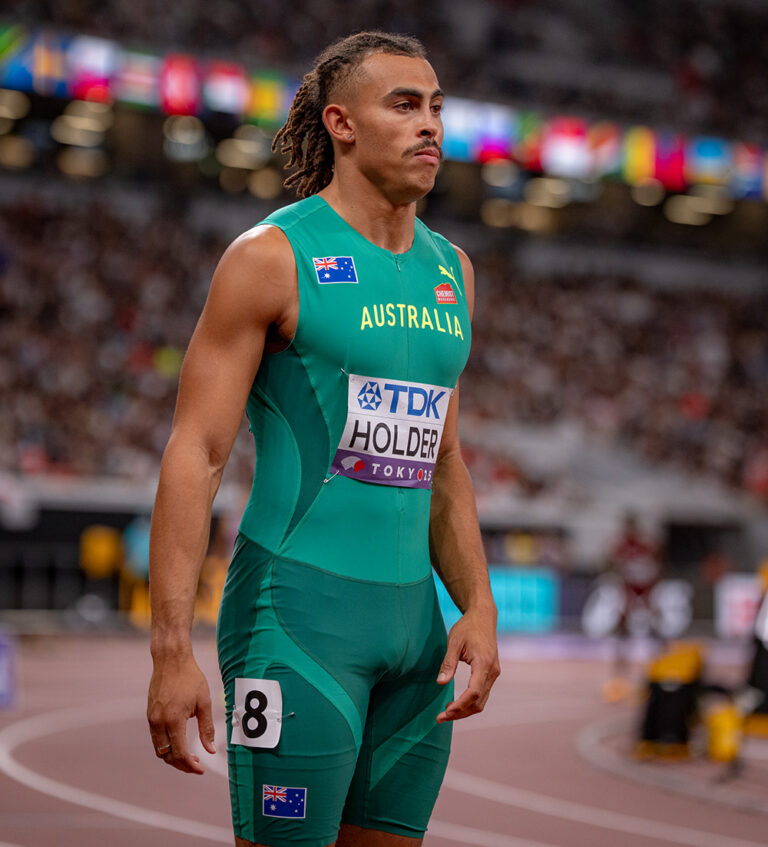 Australian sprinter in teal Australia uniform stands on a track with a bib reading 'TDK HOLDER TOKYO 2020' and a crowded stadium in the background.