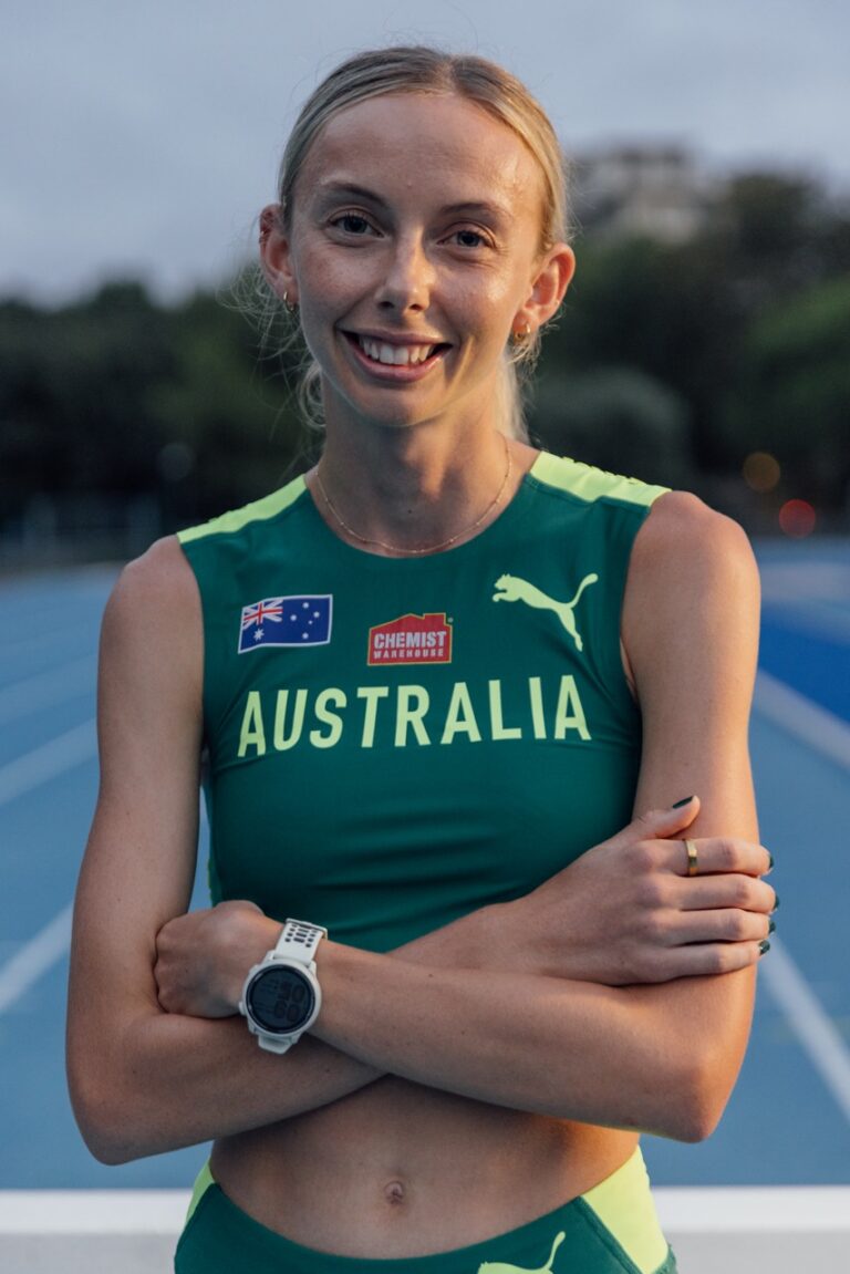Australian female sprinter in green-and-gold Australia uniform, arms crossed, smiling on a blue track.