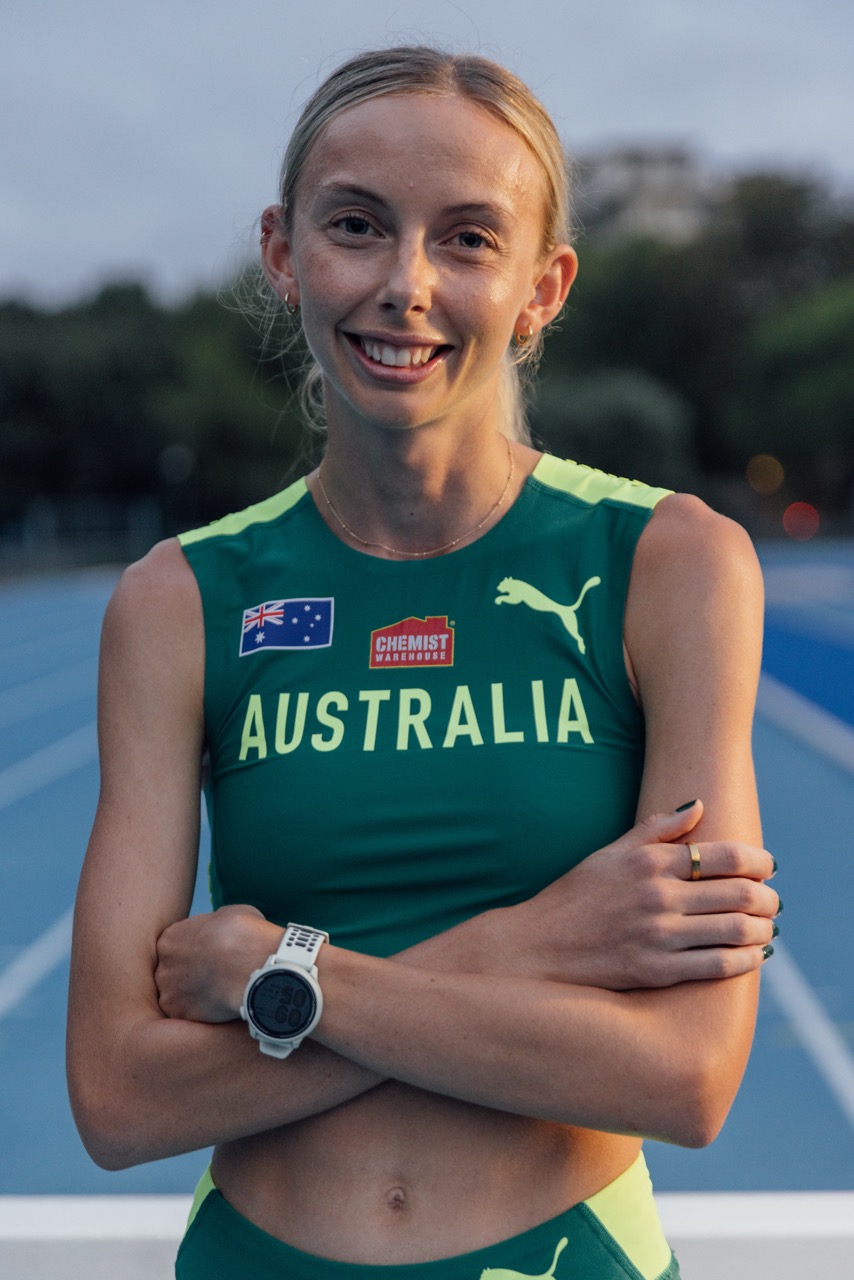 Australian female sprinter in green-and-gold Australia uniform, arms crossed, smiling on a blue track.