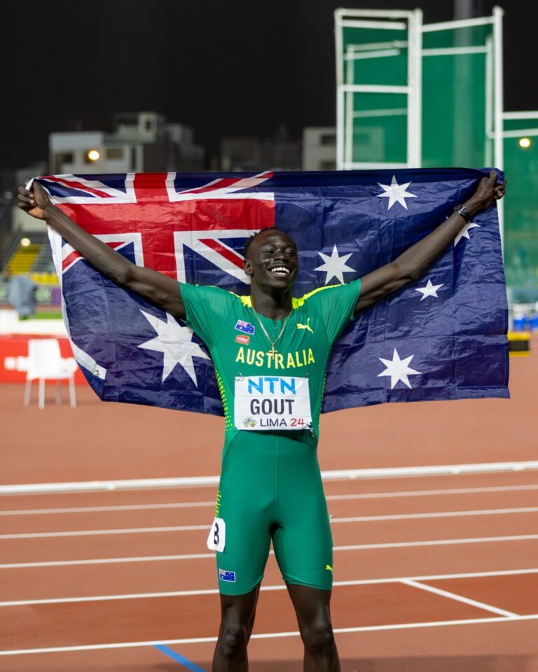 Athlete in green Australia kit holding up the Australian flag in celebration on a track field at night, arms raised in victory.