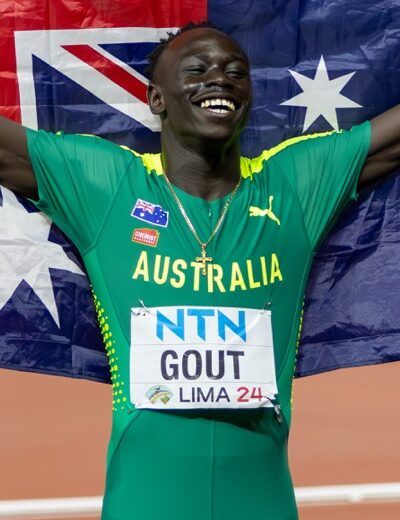 Athlete in green Australia kit holding up the Australian flag in celebration on a track field at night, arms raised in victory.