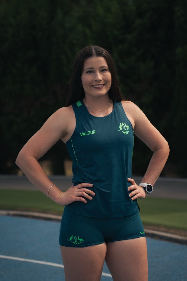 Athletic woman standing outdoors with hands on hips, wearing a teal sleeveless top marked VALOUR.