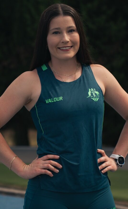 Athletic woman standing outdoors with hands on hips, wearing a teal sleeveless top marked VALOUR.