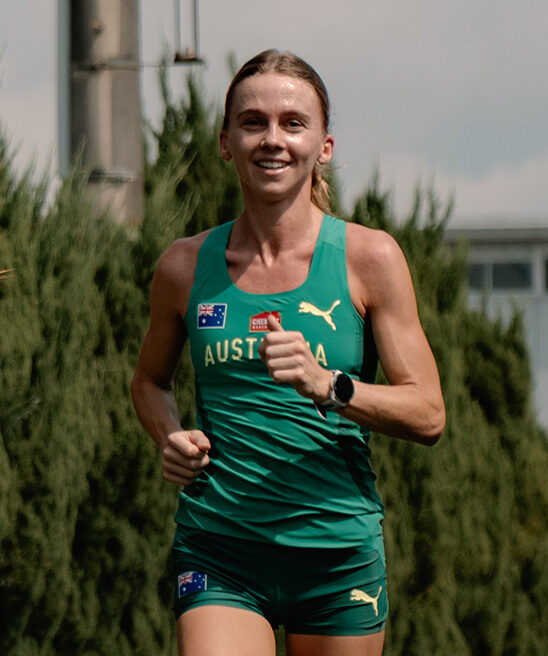 Female athlete in a teal Australia running kit smiling while jogging outdoors with trees in the background