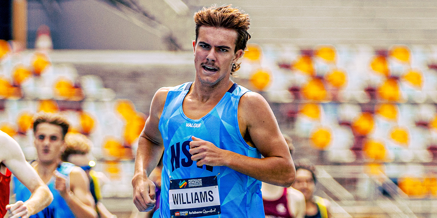 Male sprinter in a blue race singlet running in a stadium with a bib labeled WILLIAMS.