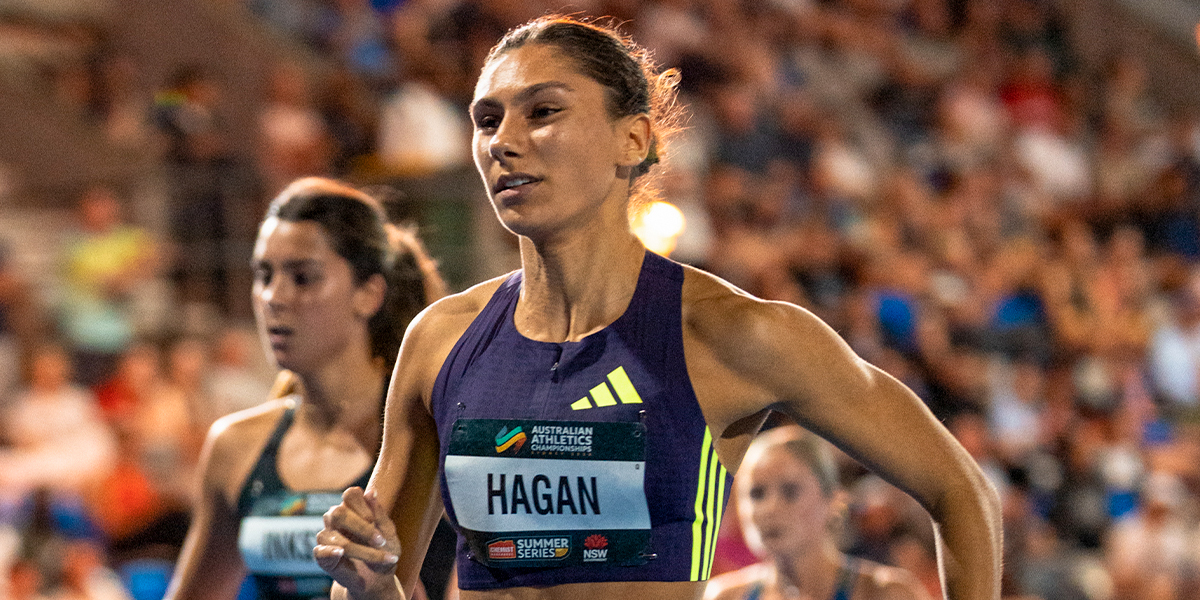 Female sprinter in a purple Adidas top with bib Hagan racing on a track with a cheering crowd in the stands.
