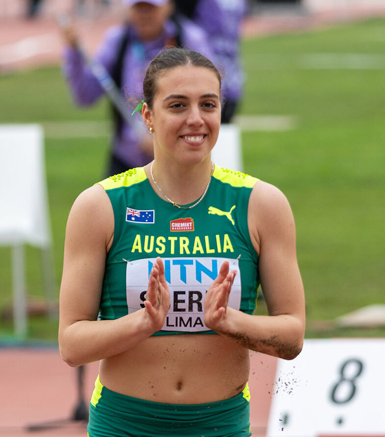 Australian female sprinter in green-and-yellow kit standing on a track, clapping with a smile after a race, spectators blurred in the background.