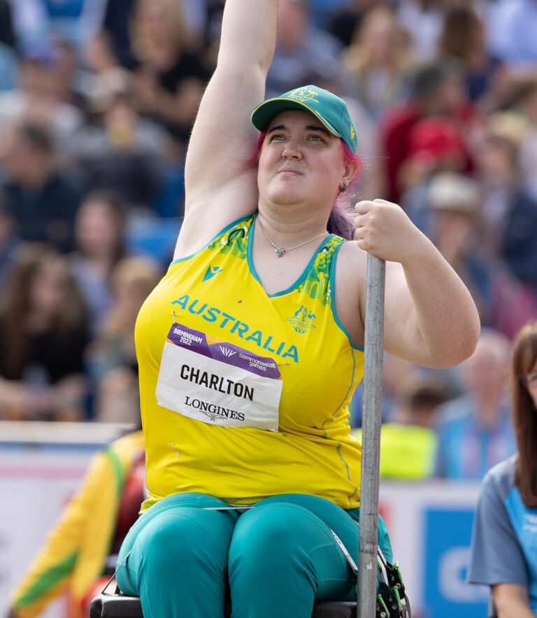 Paralympic shot put athlete in a yellow Australia jersey raises a metal ball above her head in a stadium with a cheering crowd behind her.