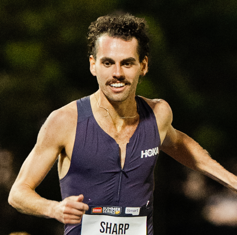 Male track runner in a sleeveless navy uniform with a bib reading SHARP, mid-race with outstretched arms.