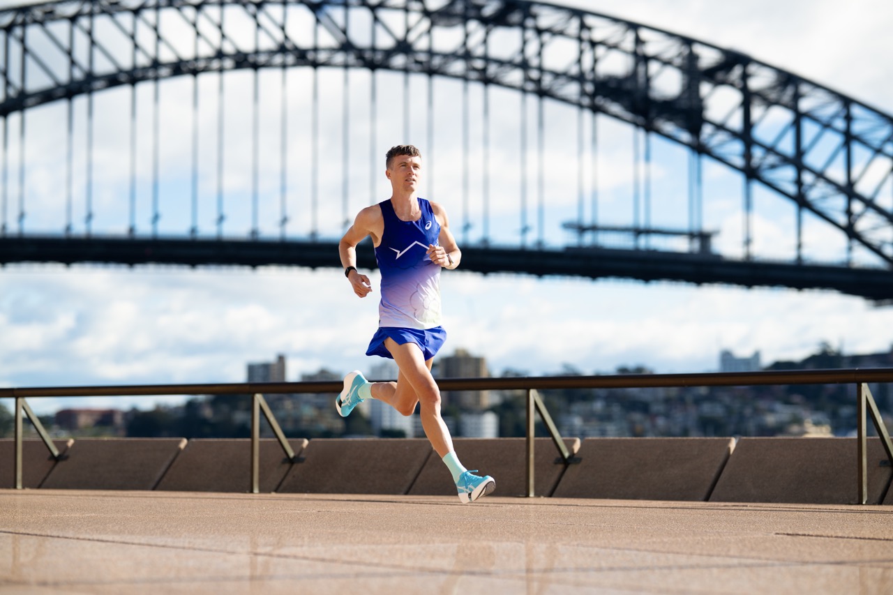 Runner in a blue gradient outfit sprinting along a riverside promenade with a large arched bridge in the background.