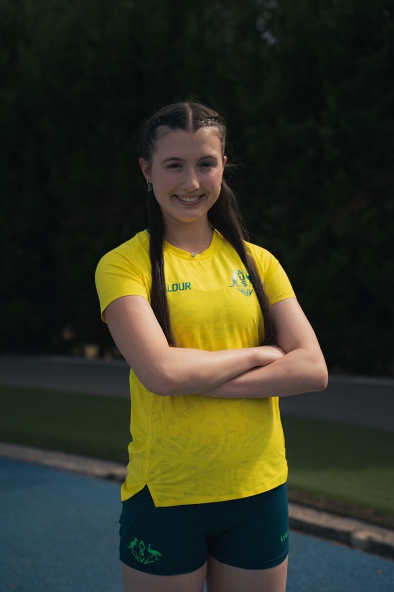 Smiling young woman in a bright yellow sports shirt and teal shorts, arms crossed, standing on a track with trees in the background.