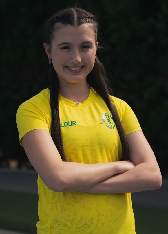 Smiling young woman in a bright yellow sports shirt and teal shorts, arms crossed, standing on a track with trees in the background.