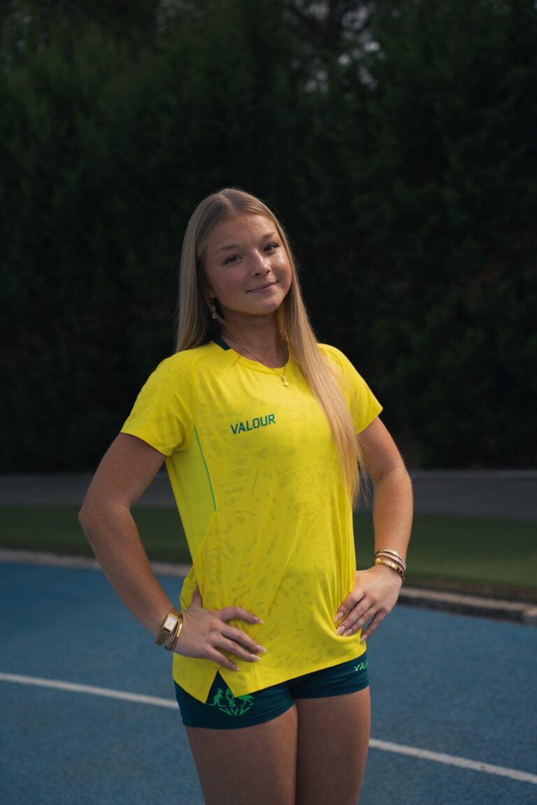 Young woman with long blond hair wearing a bright yellow VALOUR shirt, standing with hands on hips outdoors by a pool, smiling.