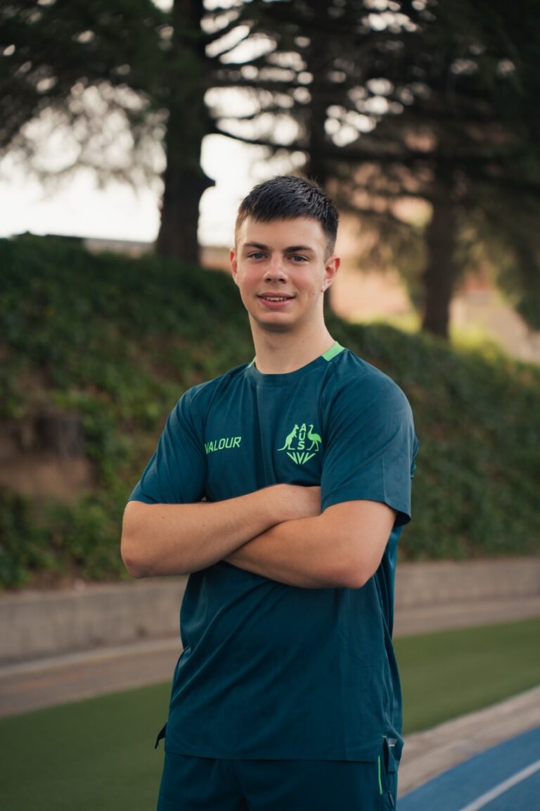 Young man in a teal athletic shirt stands outdoors with arms crossed on a track, smiling.