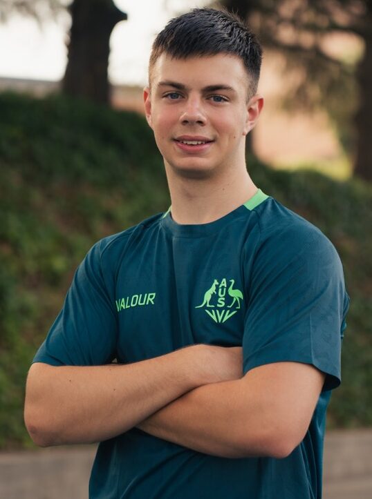 Young man in a teal athletic shirt stands outdoors with arms crossed on a track, smiling.