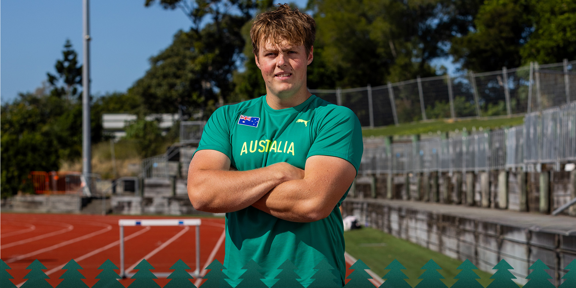 Male athlete in a teal Australia jersey stands with arms crossed on a running track outdoors, bleachers in the background.