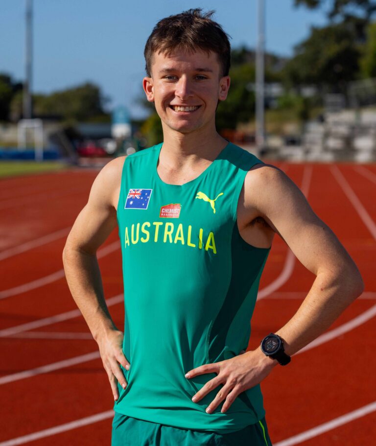 Smiling male Australian runner on a red track, teal singlet with Australian flag patch and sponsor logos, hands on hips.