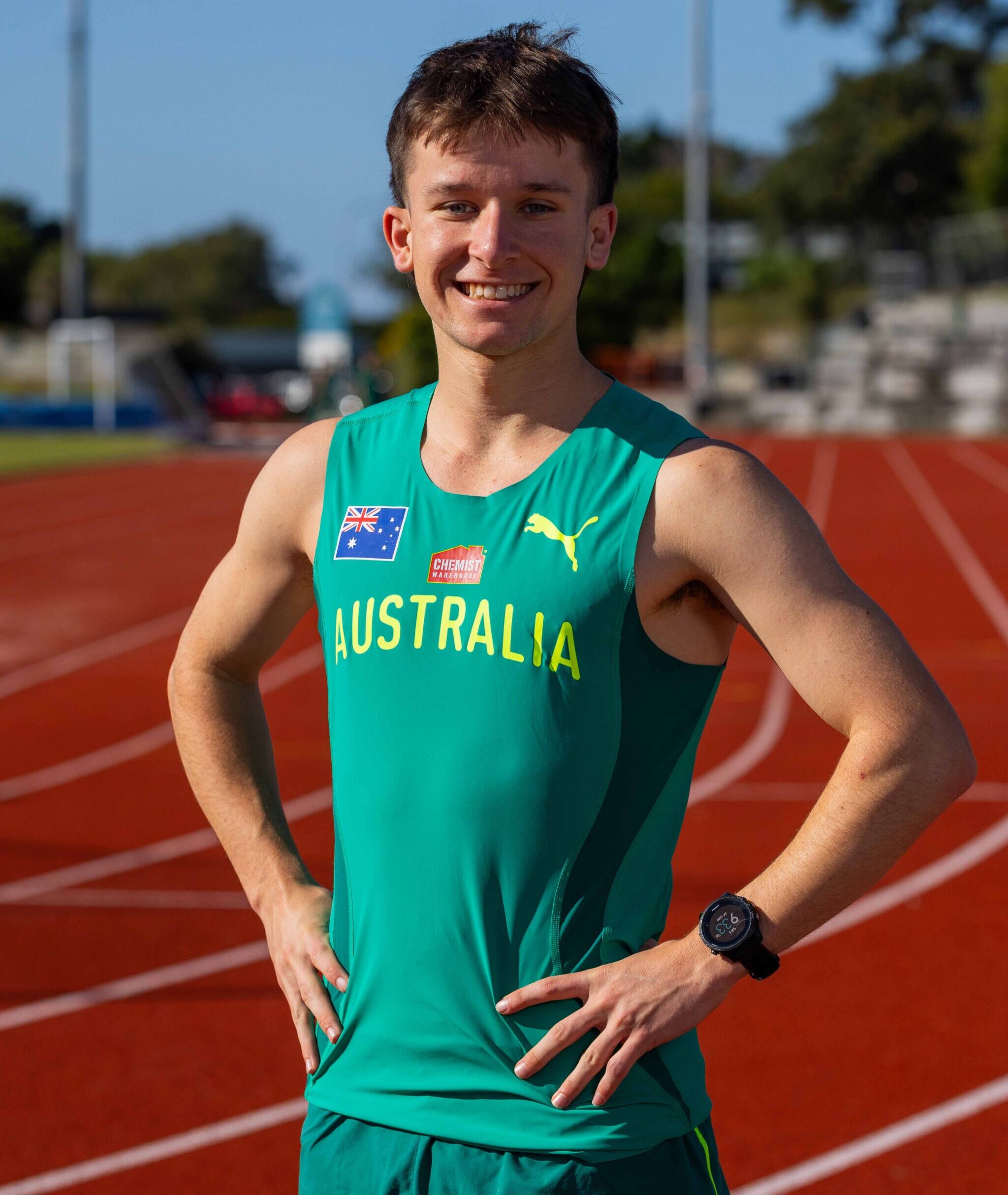 Smiling male Australian runner on a red track, teal singlet with Australian flag patch and sponsor logos, hands on hips.