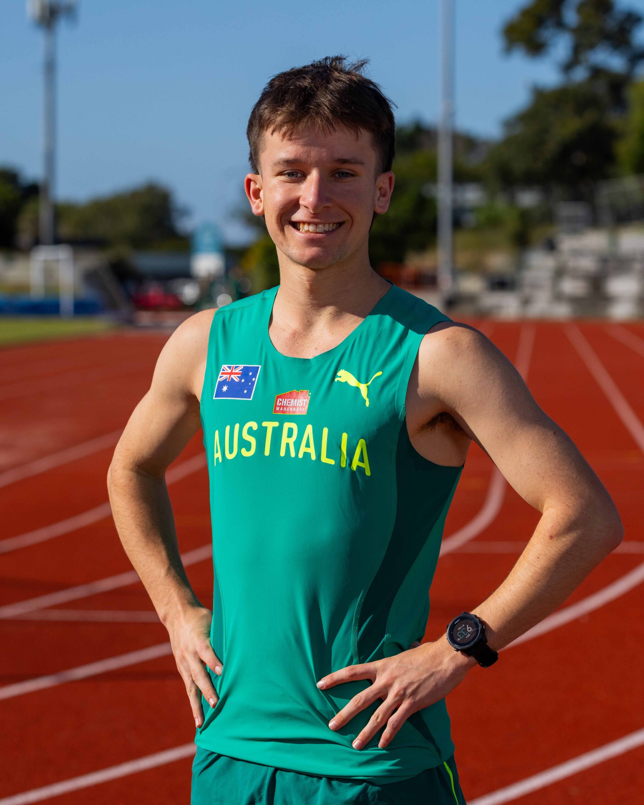Smiling male Australian runner on a red track, teal singlet with Australian flag patch and sponsor logos, hands on hips.