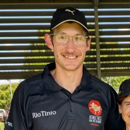 Smiling man wearing glasses and a black cap, in a dark polo with Rio Tinto and athletics logos, under a shaded structure.