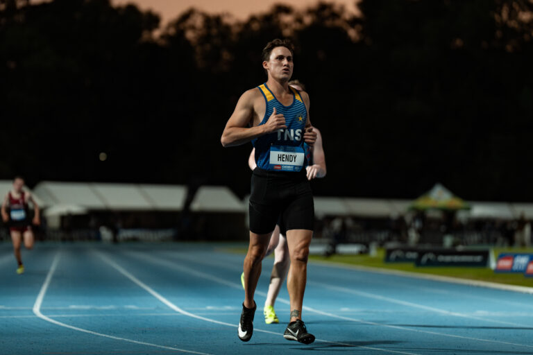 Male sprinter running on a blue track at night, wearing a blue and yellow racing singlet with bib 'HENDY'.