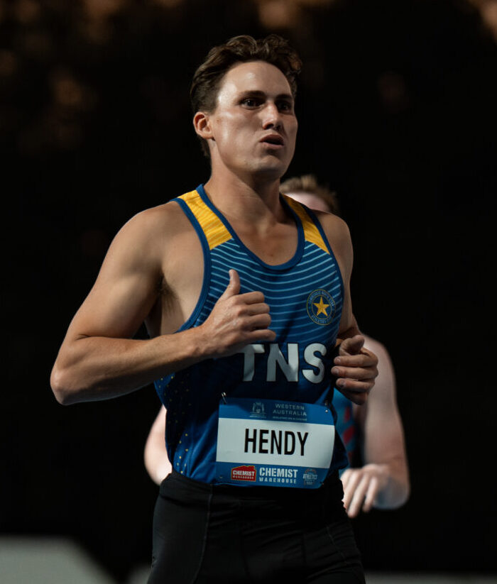 Male sprinter running on a blue track at night, wearing a blue and yellow racing singlet with bib 'HENDY'.