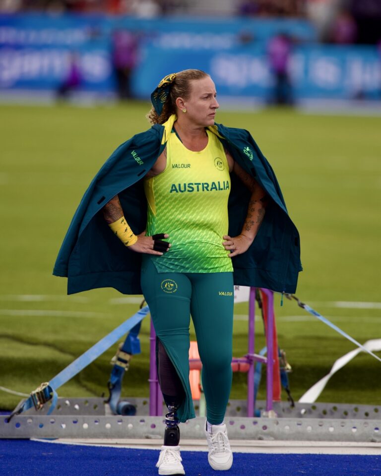Australian female para-athlete stands on track wearing a green-yellow uniform with 'AUSTRALIA' printed, a teal jacket draped over her shoulders, and a prosthetic leg visible on her left.