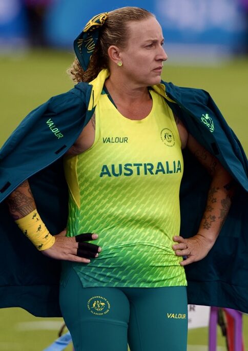 Australian female para-athlete stands on track wearing a green-yellow uniform with 'AUSTRALIA' printed, a teal jacket draped over her shoulders, and a prosthetic leg visible on her left.