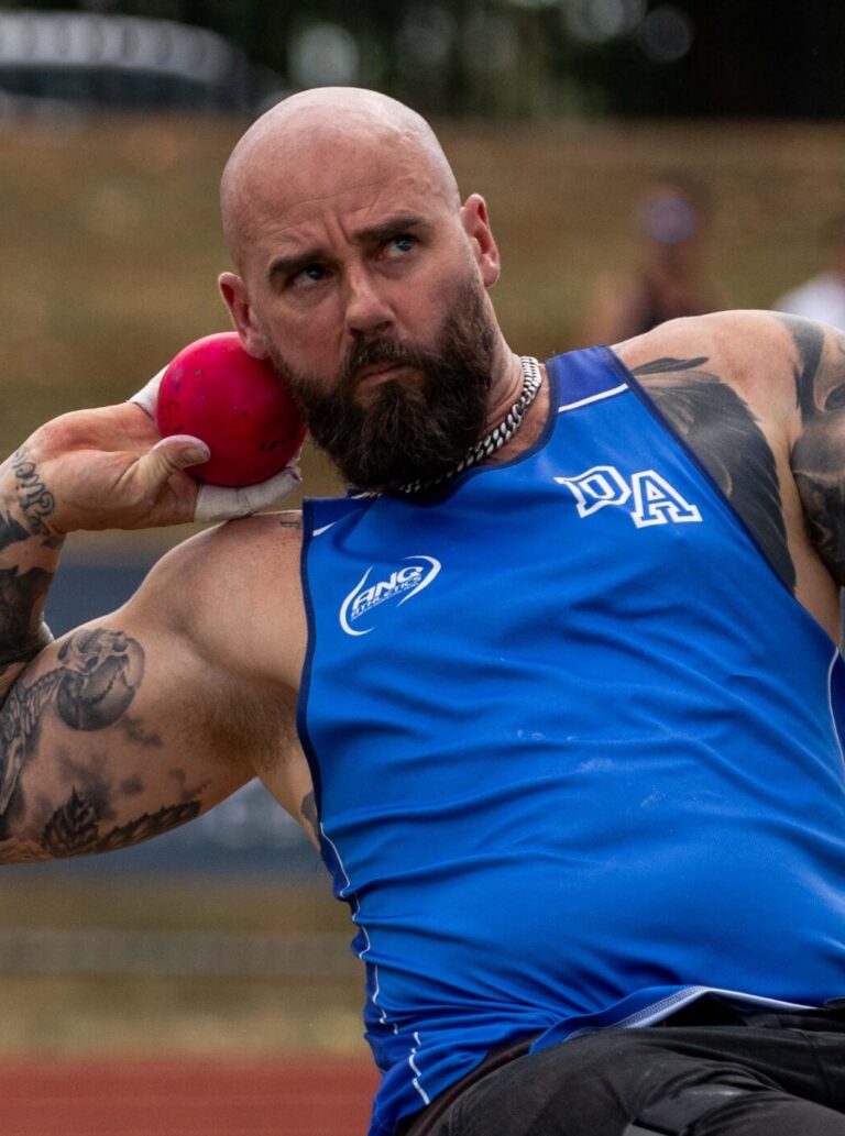 Bald muscular pole vaulter in a blue singlet gripping a pole, tattoos on his arms, ready to vault on an outdoor track with orange cones in the background