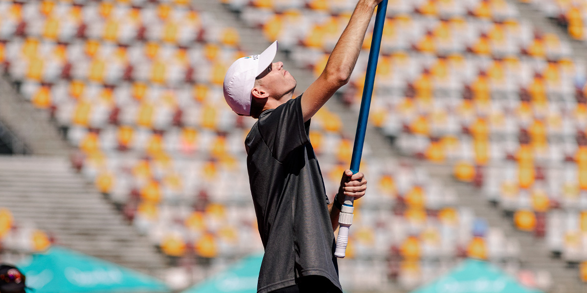 Pole vaulter in a black shirt and white cap reaches up with a blue pole in a stadium with yellow seats behind him, preparing to vault.