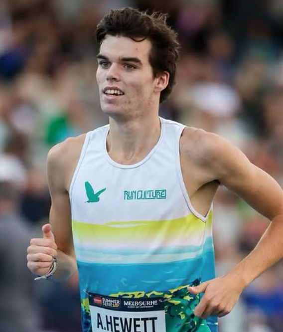 Male long-distance runner in a white-to-blue gradient singlet with a RunCulture logo, mid-race with bib A. Hewett visible in foreground.