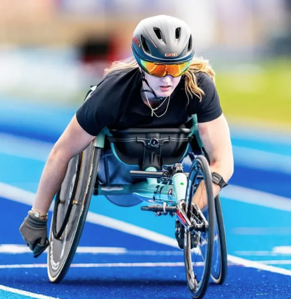 Female para-athlete in a racing wheelchair on a blue track, wearing a helmet and orange-tinted visor, leaning forward in motion.