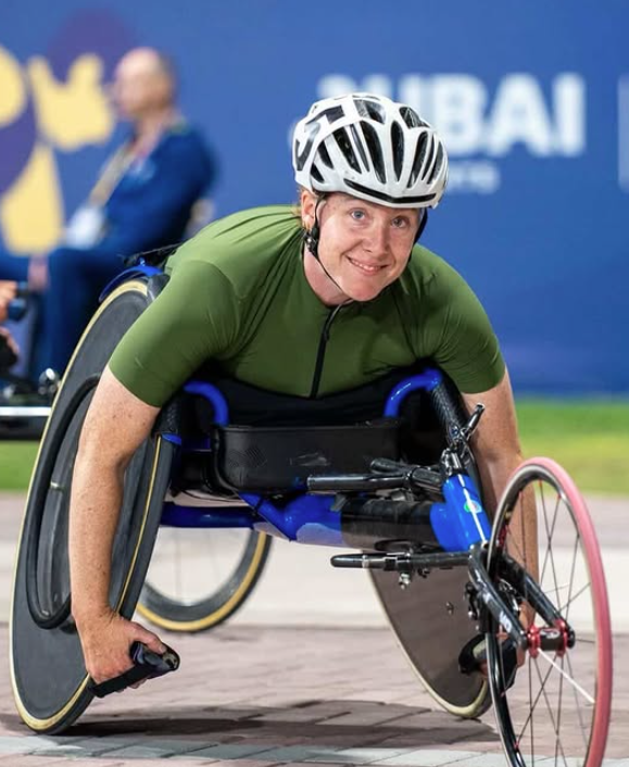 Paralympic wheelchair racer in a green jersey and white helmet, leaning forward during a race in a blue surroundings.