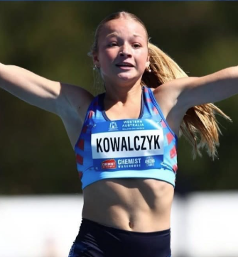Female athlete with arms raised in celebration, wearing a blue sports top and a bib reading KOWALCZYK.