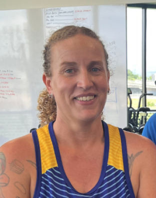 Smiling woman in a blue and yellow athletic top, with tattoos on her arms, indoors near a window.