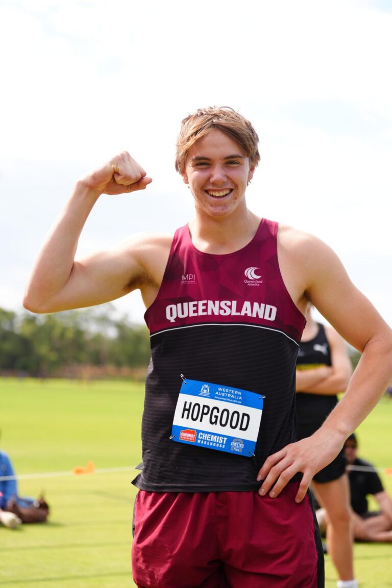 Smiling male runner in a maroon Queensland singlet flexing his right arm outdoors; bib reads HOPGOOD.