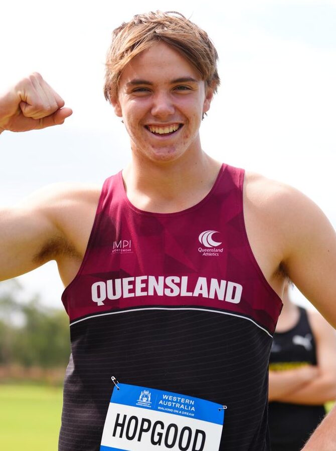 Smiling male runner in a maroon Queensland singlet flexing his right arm outdoors; bib reads HOPGOOD.