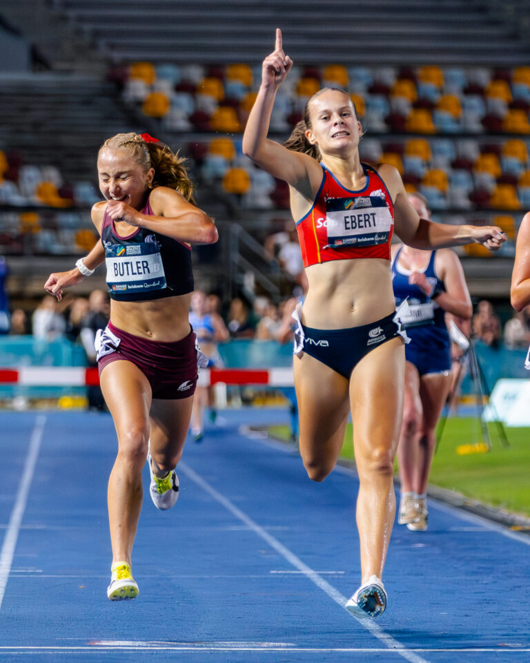 Two female sprinters near the finish line on a blue track, one raising a finger in victory as they run side by side in a stadium.