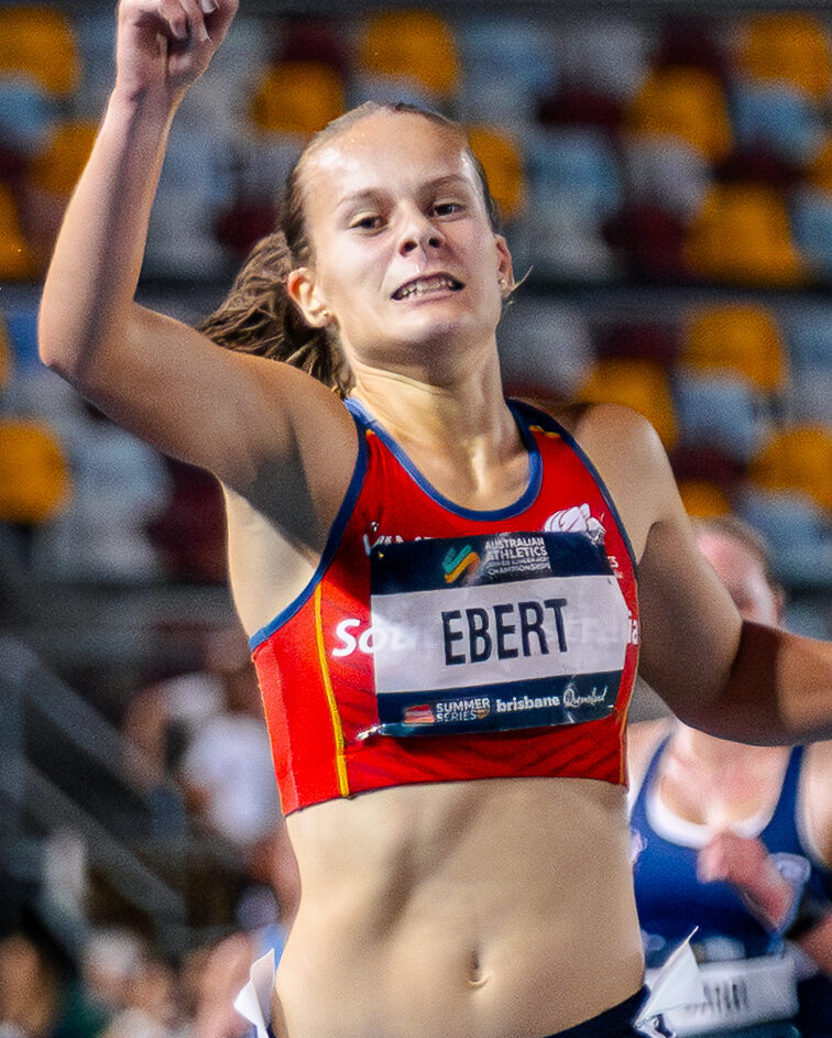 Two female sprinters near the finish line on a blue track, one raising a finger in victory as they run side by side in a stadium.