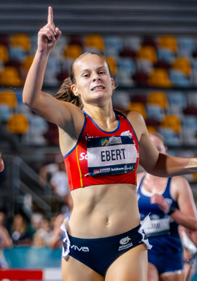 Two female sprinters near the finish line on a blue track, one raising a finger in victory as they run side by side in a stadium.