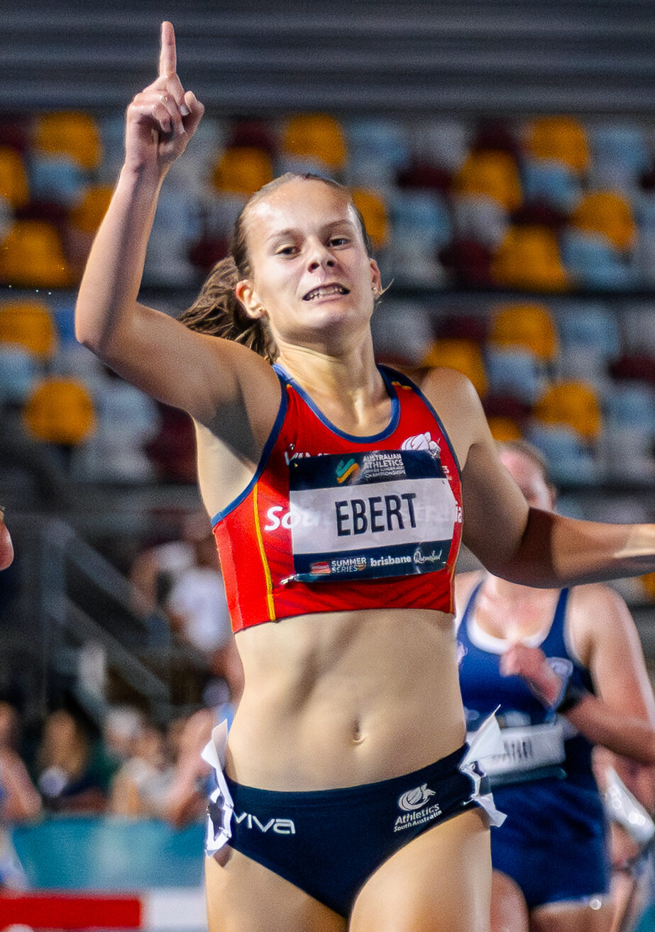 Two female sprinters near the finish line on a blue track, one raising a finger in victory as they run side by side in a stadium.