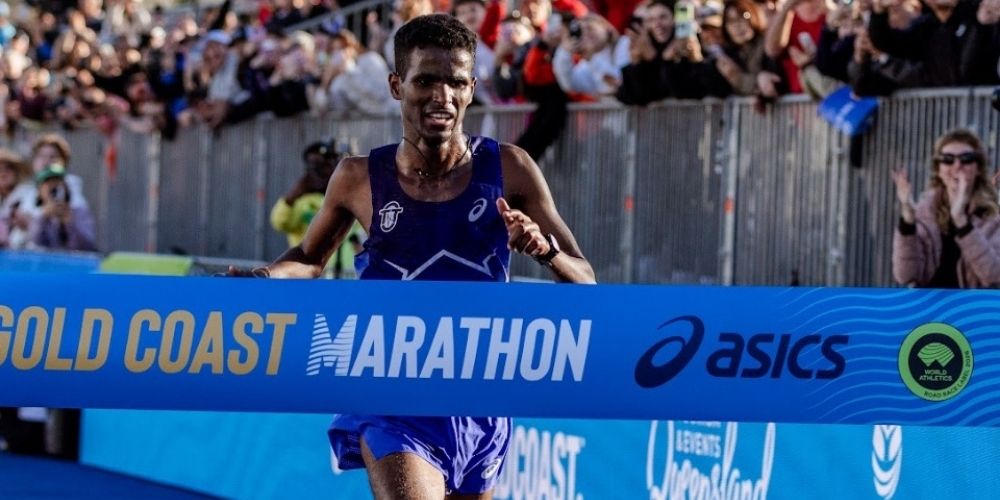 Male runner in blue kit crosses the finish line at the Gold Coast Marathon as spectators cheer behind a branded blue banner.