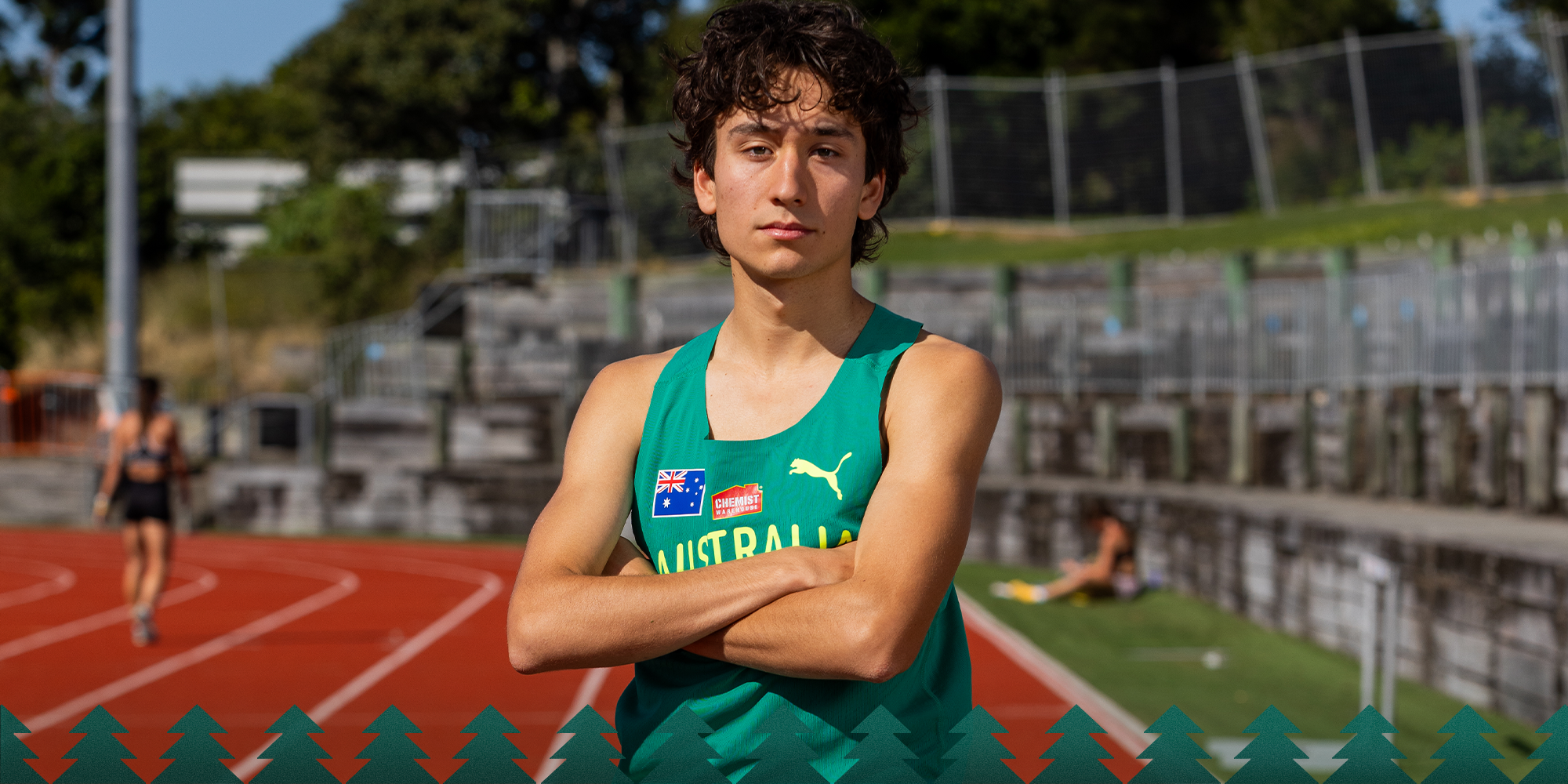 Young male athlete wearing Australia's green team singlet stands on a running track with arms crossed.