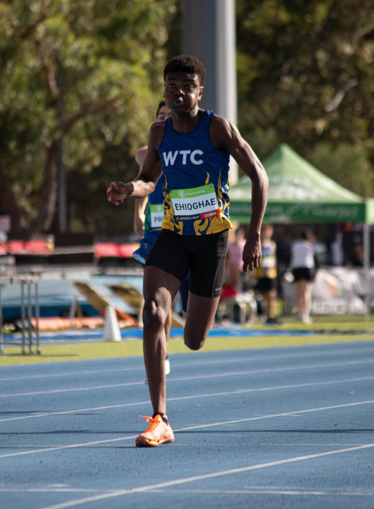 Male sprinter in a blue WTC singlet racing, bib shows EHIOGHAE on his chest