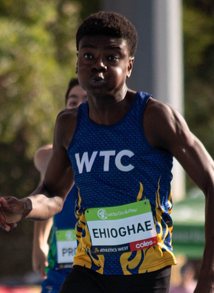 Male sprinter in a blue WTC singlet racing, bib shows EHIOGHAE on his chest
