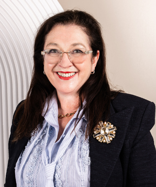 Smiling woman in a dark blazer with a decorative brooch, wearing glasses and a striped blouse, posing for a portrait.
