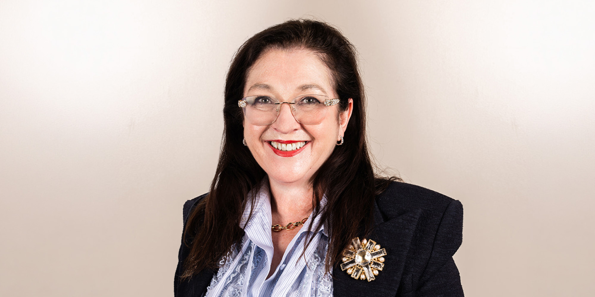 Professional portrait of a smiling woman with dark hair and glasses, wearing a navy blazer, striped scarf, and gold brooch.