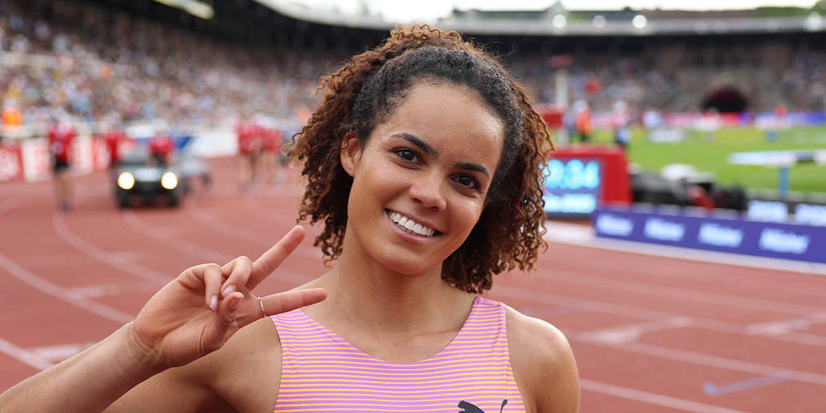 Smiling female sprinter on a track flashing a peace sign with blurred stadium stands behind her.